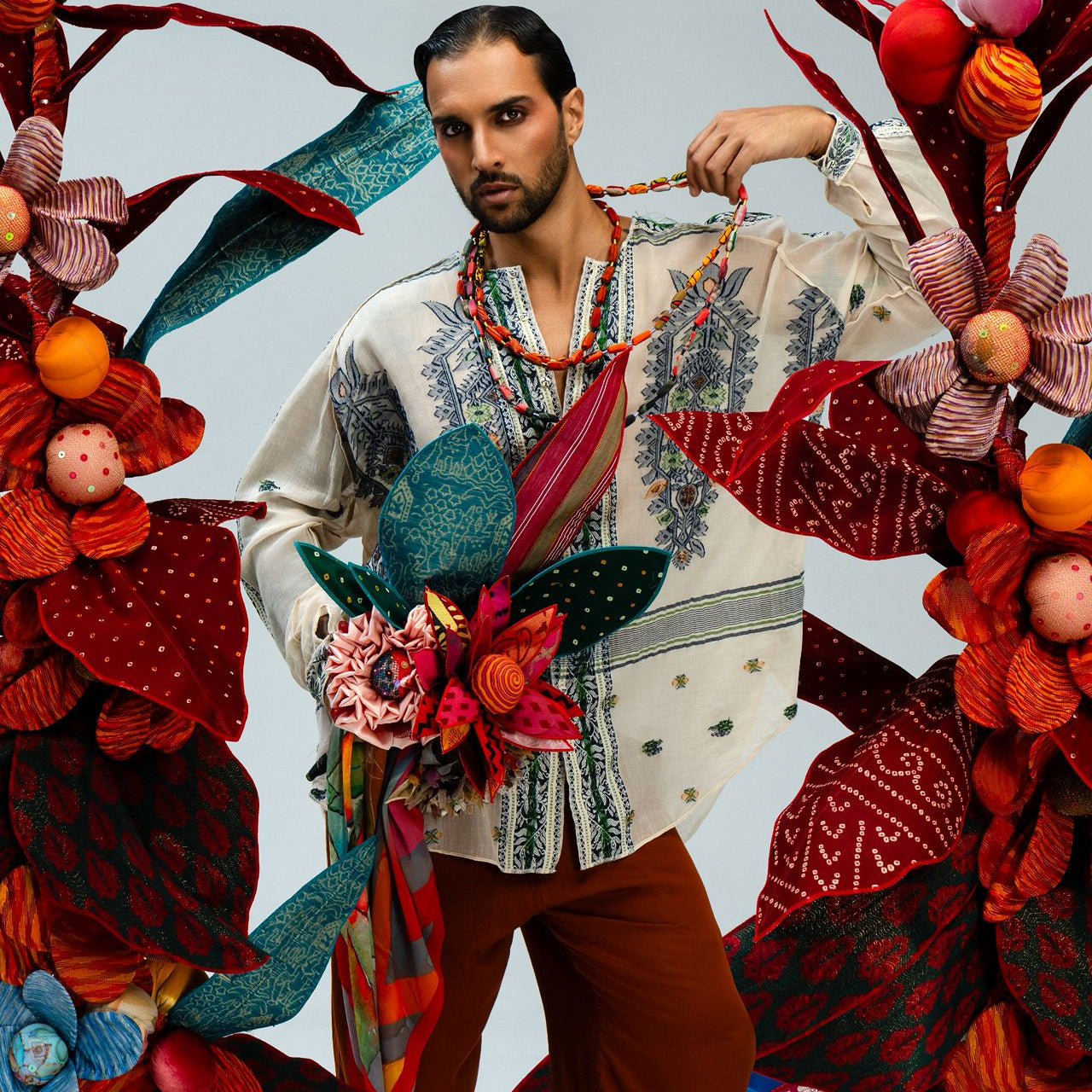 Bohemian style editorial fashion shot featuring model in PAUL ROPP's multi-color layered beaded necklaces, patterned bandeau top in broken white color, and red longpants against a fabric floral backdrop.